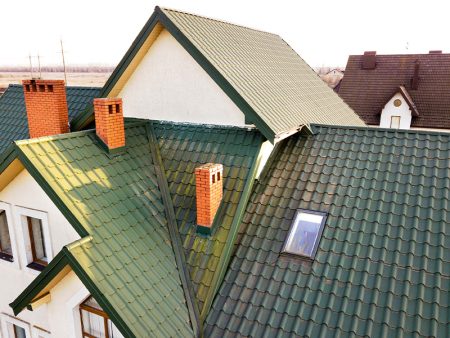 Green metal shingled house roof with attic plastic window and brick chimney.